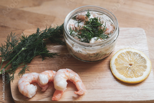 Scandinavian Food - Wooden chopping board with fresh shrimps, dill, slice of lemon and glass jar of traditional Skagen 