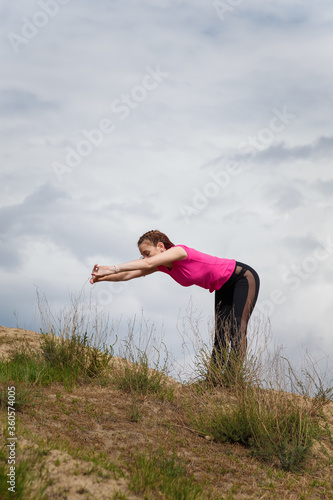 Young sporty woman in sportswear on hill top stretching or warming up on cloudy sky background. Fitness, healthy way of life, wellbeing, freedom, loosing weight concept.
