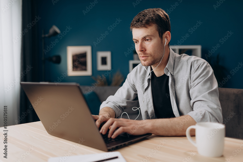 Young man with light stubble sitting at table in earphones and working on laptop. Company worker doing his job on distance while staying at home. Young man with light stubble sitting at table in earphones and working on laptop. Company worker doing his job on distance while staying at home.