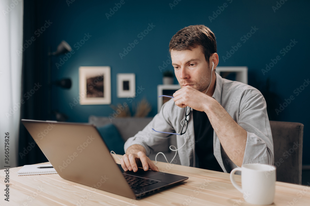 Concentrated bearded man in casual clothing using laptop for remote work at home. Young guy in earphones working on computer while sitting at table. Concentrated bearded man in casual clothing using laptop for remote work at home. Young guy in earphones working on computer while sitting at table.
