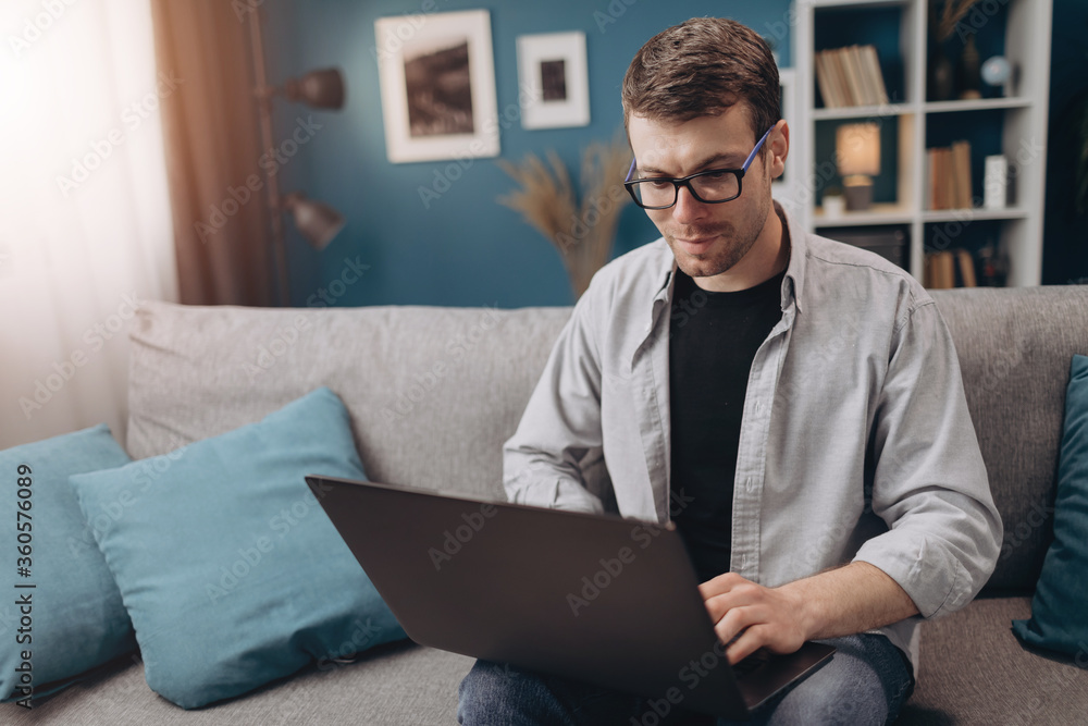 Happy young male in casual outfit sitting on comfortable couch and using portable laptop. Bearded guy in eyeglasses relaxing at home with modern gadget Happy young male in casual outfit sitting on comfortable couch and using portable laptop. Bearded guy in eyeglasses relaxing at home with modern gadget
