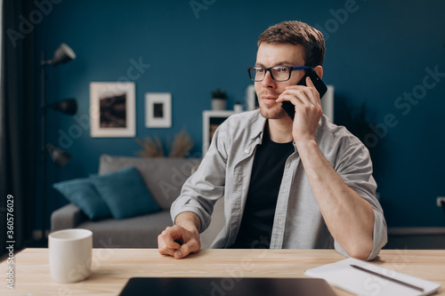 Handsome guy in casual outfit sitting at table and talking on mobile phone. Young man in eyeglasses using smartphone for conversation while staying at home.