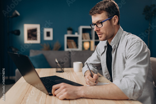 Side view of bearded man in eyeglasses and casual shirt making notes while working on laptop at home. Young guy sitting at table and using modern device.