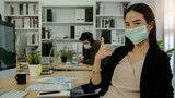 business woman wearing mask sitting in office and show thumb up