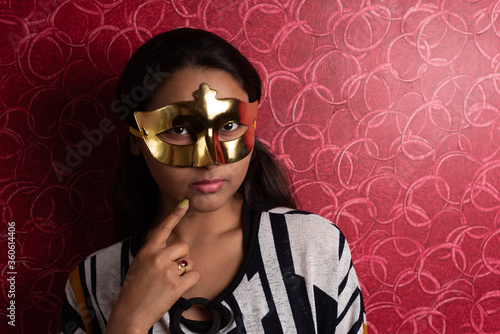 Fashion portrait of an Indian Bengali beautiful and young girl in western dress wearing an eye mask standing in front of a red textured wall. Indian fashion and portrait