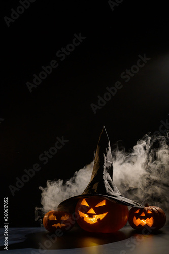 Vertical Halloween card. Witch hat on a pumpkin with carved creepy grimaces on a black background in the fog. Jack-o-lantern glows from the inside and emits smoke.