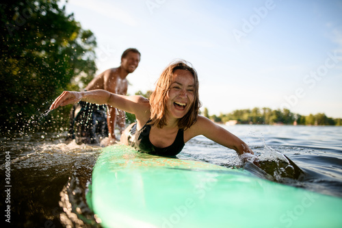 Tableau sur toile laughing woman lies on surfboard and rows her hands and male instructor supports