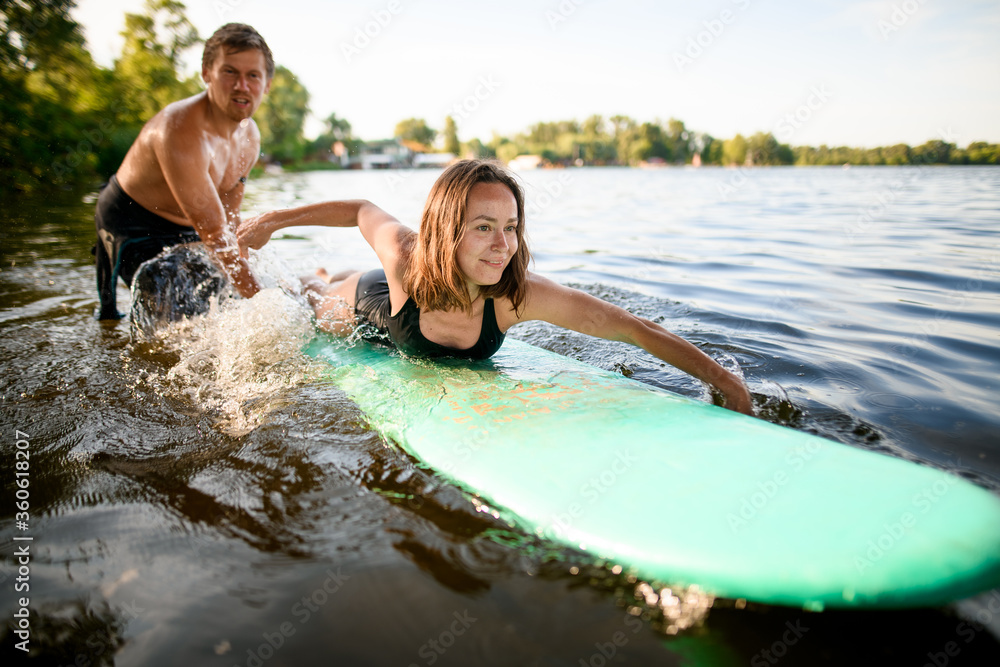 View on woman who lies on surfboard and rows her hands and male instructor supports her View on woman who lies on surfboard and rows her hands and male instructor supports her