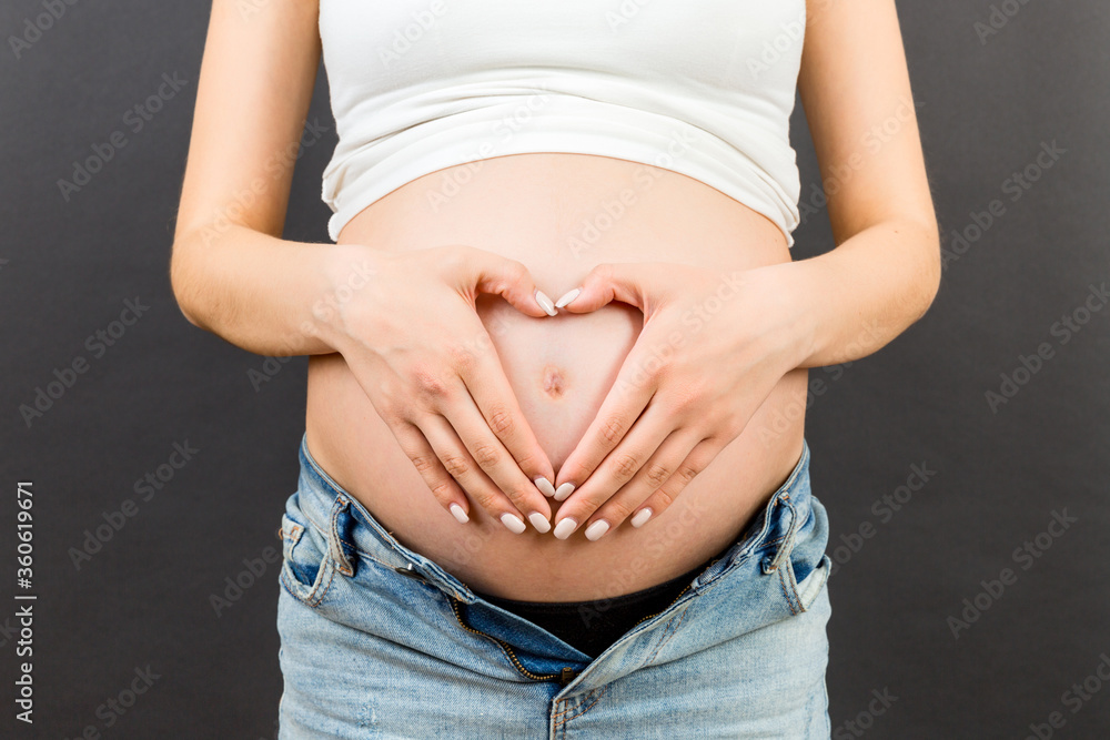 Close up of pregnant woman in opened jeans making heart on her belly at colorful background with copy space. Love to a future baby concept Close up of pregnant woman in opened jeans making heart on her belly at colorful background with copy space. Love to a future baby concept
