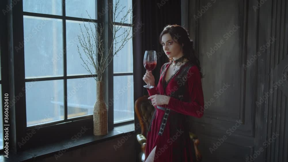 Beautiful young woman queen holding glass bloody wine in hands. Lady Brunette long wavy hair. Red sexy carnival vampire dress costume. dark black interior gothic medieval room, window. Fashion model Beautiful young woman queen holding glass bloody wine in hands. Lady Brunette long wavy hair. Red sexy carnival vampire dress costume. dark black interior gothic medieval room, window. Fashion model