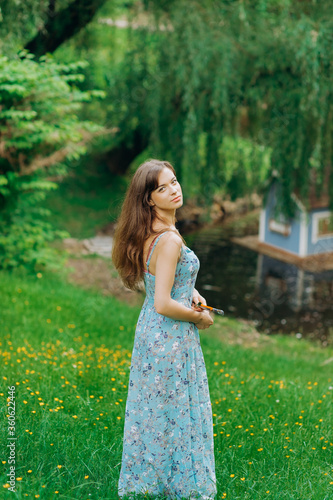 Portrait of a young artist in a blue dress with a print in the park near the lake with brushes in her hand © Yuliia