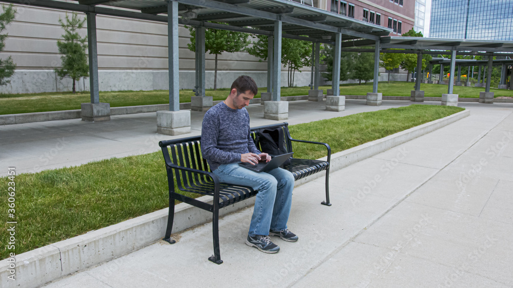 High quality picture of a young men who is sitting on a train station and working on his laptop which is holding on the lap. Young men sitting on a black metal banch on a station in the city. High quality picture of a young men who is sitting on a train station and working on his laptop which is holding on the lap. Young men sitting on a black metal banch on a station in the city.