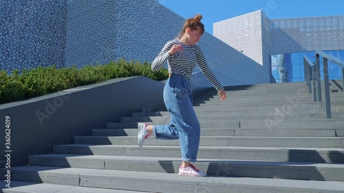 A happy teenage girl is dancing a shuffle on the stairs and enjoying life next to a modern building. The concept of carefree, youth culture and city life