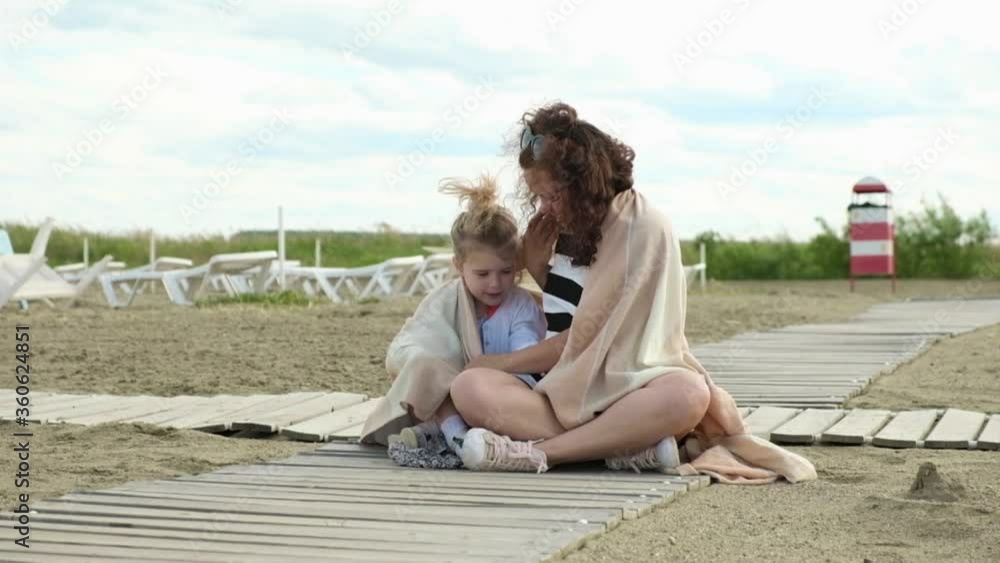 mother and daughter sitting on the sand hugging and wrapped in a warm blanket from the wind. Family weekend active at the sea. Outdoor activities on weekends. A family on a sandy beach is resting. mother and daughter sitting on the sand hugging and wrapped in a warm blanket from the wind. Family weekend active at the sea. Outdoor activities on weekends. A family on a sandy beach is resting.