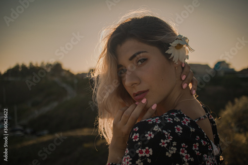young curly blonde in a dark floral blouse with a camomile behind her ear against the sky at sunset