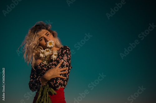 young curly blonde with a camomile in her mouth and daisies in her hands against the sky