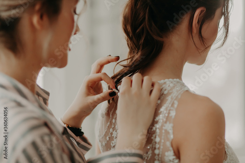 bridesmaid helps fasten wedding dress in room, preparing for ceremony