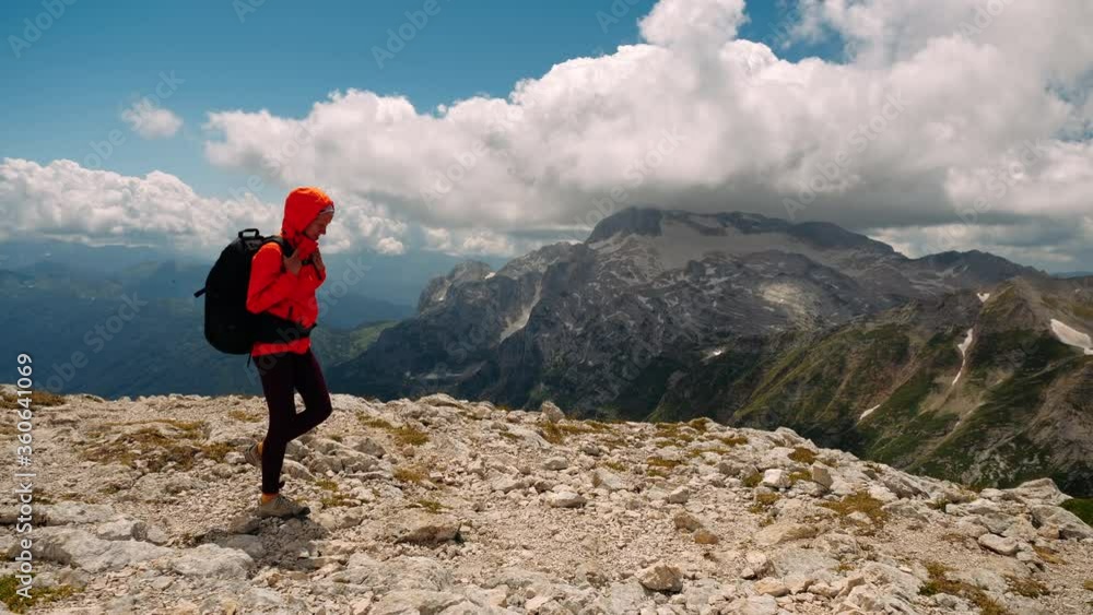 Side view of active female tourist with backpack walking along edge of high mountain and going down after conquering peak, background of beautiful mountains and clouds. Hiking and healthy lifestyle. Side view of active female tourist with backpack walking along edge of high mountain and going down after conquering peak, background of beautiful mountains and clouds. Hiking and healthy lifestyle.