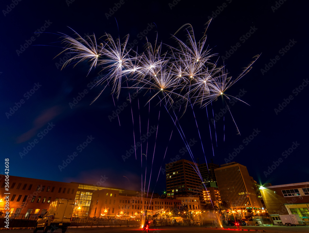 Fireworks. Wide angle shot showing the launch as well as the blooms. Buildings and street lights in the background. Focus is on the fireworks.