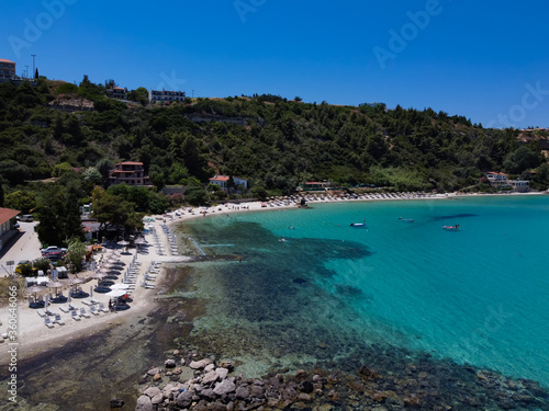 Mediterranean terrain Greek beach landscape drone shot. Aerial top view of Afytos or Athitos hilltop village at Kassandra Chalkidiki peninsula with green plantation by sandy beach with umbrellas.