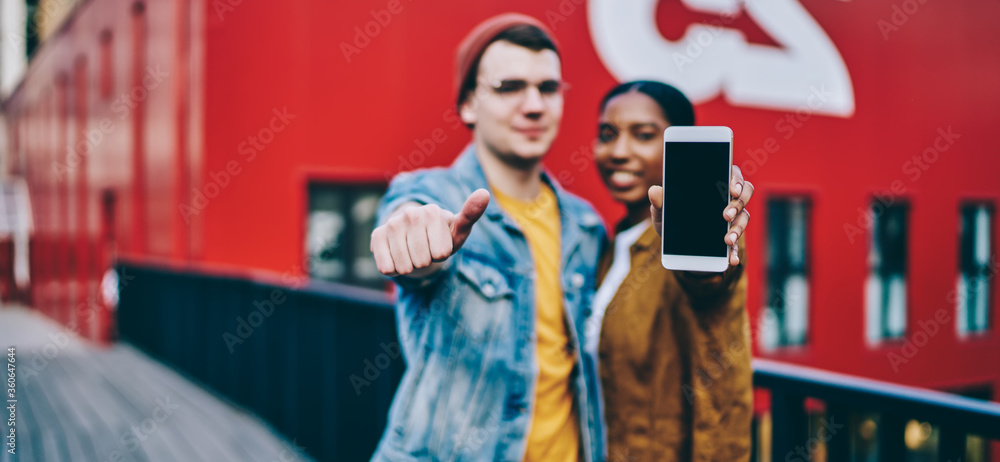 Obraz premium Multiracial hipster guys showing cellphone device with copy space area for internet advertising, blurred millennial male and female promote mock up mobile phone with blank screen for your website