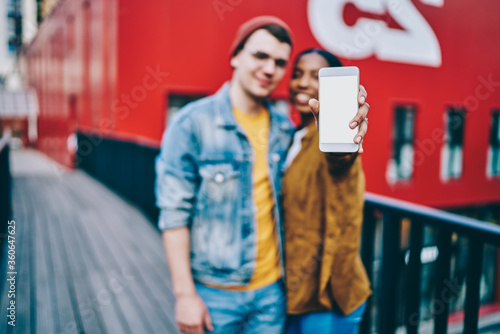 Selective focus on modern mobile phone with template mock up screen, blurred male and female millennial generation hipster couple standing outdoors holding smartphone with new application, copy space