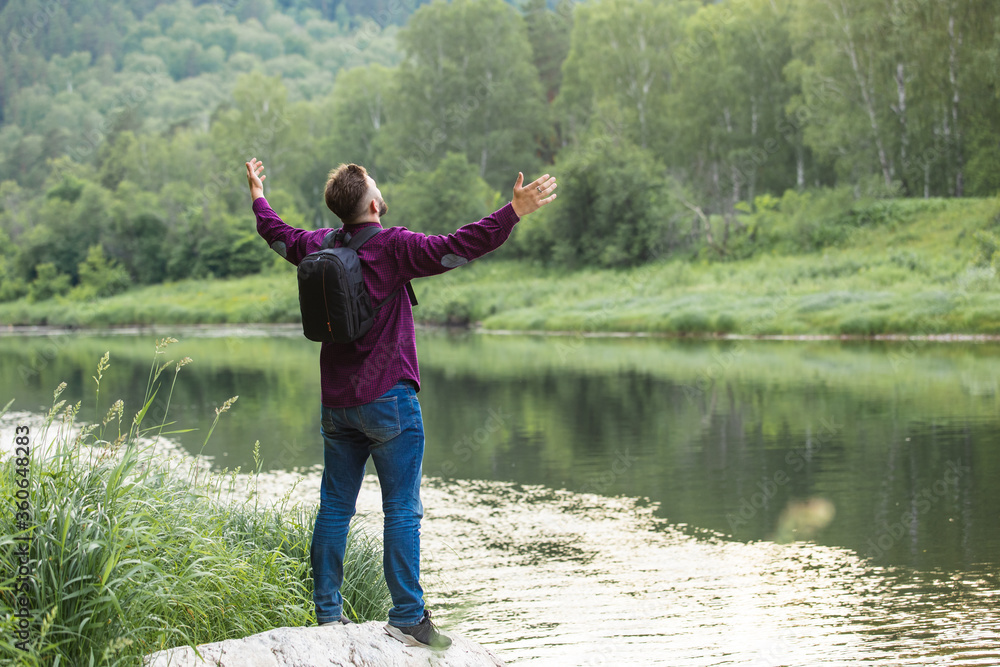
The guy - a tourist on the river bank opened his hands to the sides, raised his hands up. The man’s position is behind, in full growth, surrounded by nature