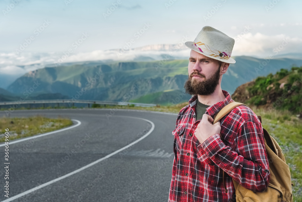 Handsome young man in a straw hat and a plaid shirt, stands on the road, against the backdrop of beautiful mountains at sunset. The concept of freedom and hitchhiking Handsome young man in a straw hat and a plaid shirt, stands on the road, against the backdrop of beautiful mountains at sunset. The concept of freedom and hitchhiking