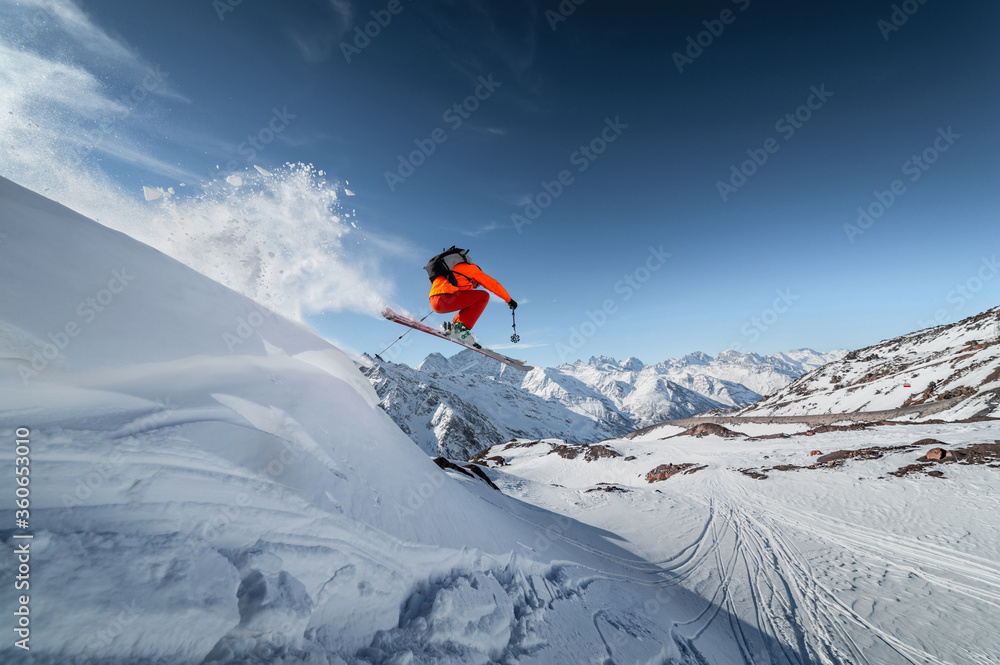 Athlete male skier jumps from a snow-covered slope against the backdrop of a mountain landscape of snow-covered mountains on a sunny day. The concept of winter sports wide angle