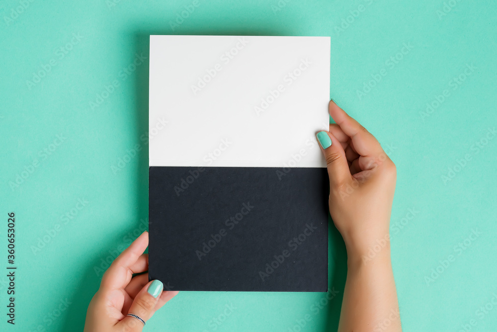Female's hands are holding empty mockup duotone white black paper sheet for text or message above pastel turquoise background. Female's hands are holding empty mockup duotone white black paper sheet for text or message above pastel turquoise background.