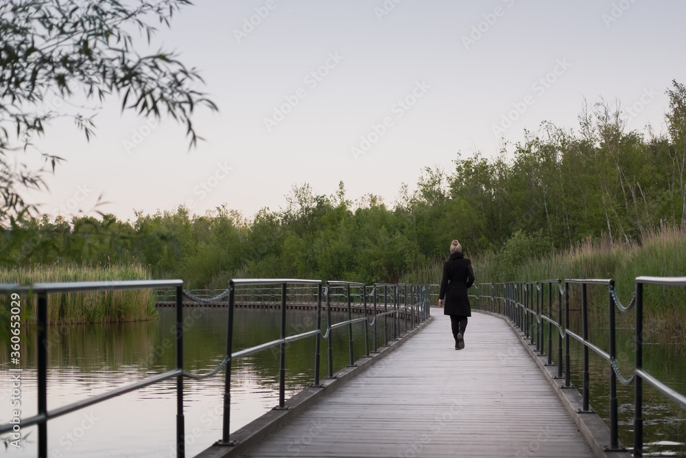Blonde woman in black coat walking on a bridge over the clam lake in the evening