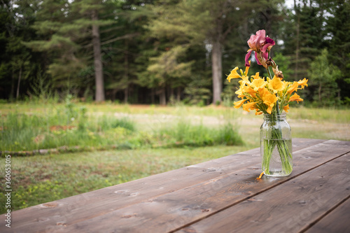 Bouquet of yellow and purple flowers on a picnic table in a garden setting. Everything is wet from recent rain. photo