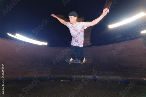 A young skateboarder rides at speed sideways along the ramp by spreading his hands to the sides. Night shot with long exposure at night speed in a skatepark