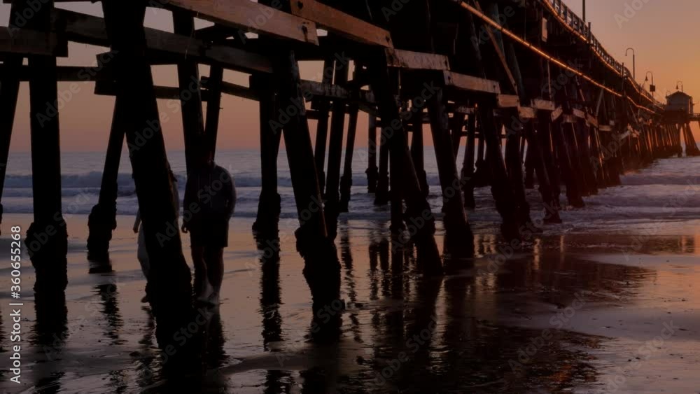 Couple walking holding hands together at Scenic orange pink Sunset with epic rays of light and sun flare under wooden pier in San Clementa California Couple walking holding hands together at Scenic orange pink Sunset with epic rays of light and sun flare under wooden pier in San Clementa California