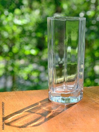 Empty drink glass against a background of unfocused green foliage with a shadow from the sun's rays
