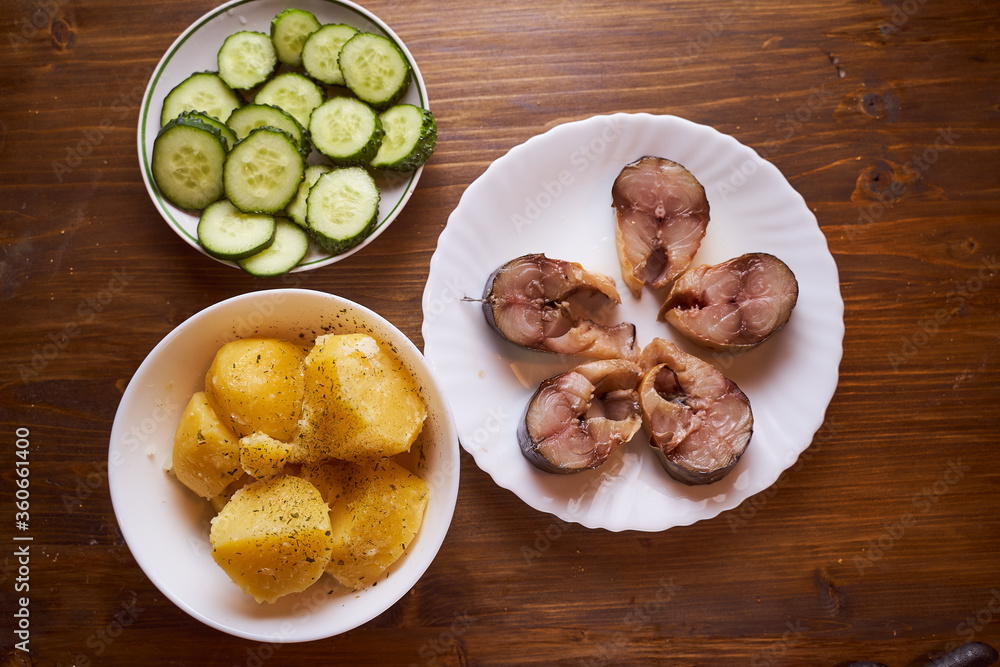 Boiled potatoes, cucumbers and sardines in plates on a brown table