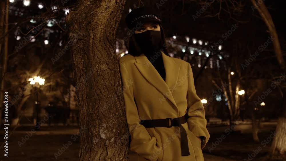 Nice brunette girl with her hair in a black virus mask in a beige coat is standing by a tree in a park in the night city and looks around slowmotion Nice brunette girl with her hair in a black virus mask in a beige coat is standing by a tree in a park in the night city and looks around slowmotion