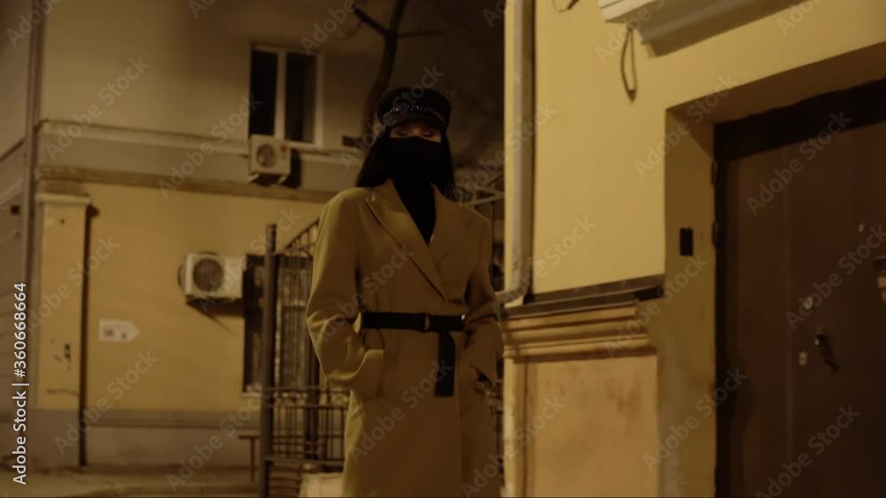 pretty brunette girl with her hair in a black virus mask in a beige coat walks along building in the night city hands in pockets pretty brunette girl with her hair in a black virus mask in a beige coat walks along building in the night city hands in pockets