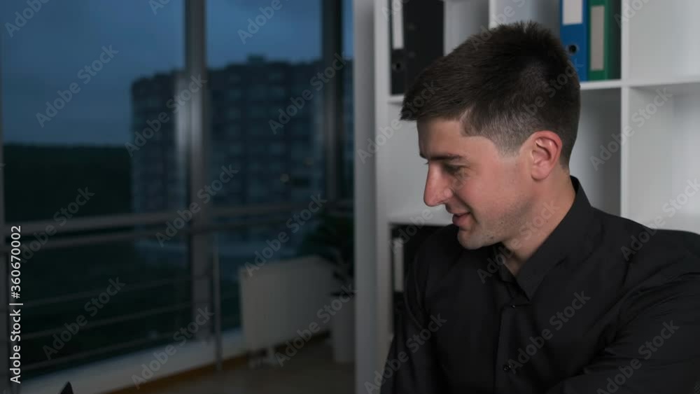 Young handsome man of Caucasian appearance working at the table typing text on laptop solves work issues. Managed at night Young handsome man of Caucasian appearance working at the table typing text on laptop solves work issues. Managed at night