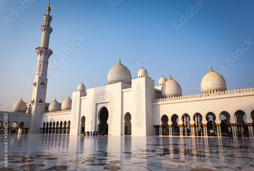 The entrance of Grand Mosque in Abu Dhabi during sunset time. People are at the entrance and willing to go inside the mosque for praying. Minaret and domes are seen.