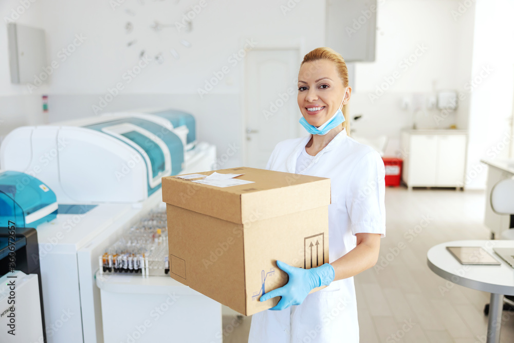 Smiling blond female lab assistant standing and holding box with medicines and vaccine. Smiling blond female lab assistant standing and holding box with medicines and vaccine.