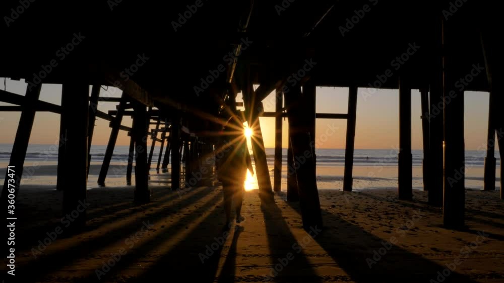 Woman silhouette walking under pier at Scenic orange pink Sunset with epic rays of light and sun flare wooden pier in San Clementa California Woman silhouette walking under pier at Scenic orange pink Sunset with epic rays of light and sun flare wooden pier in San Clementa California