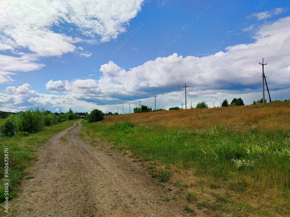 country road along the power line among green and red grass in a field against a blue sky with unusual clouds