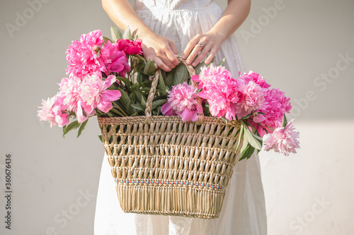 Fototapeta Naklejka Na Ścianę i Meble -  Boho girl holding pink peonies in straw basket. Stylish hipster woman in bohemian floral dress gathering peony flowers on white backgrounds. International Womens Day. Wedding decor and arrangement
