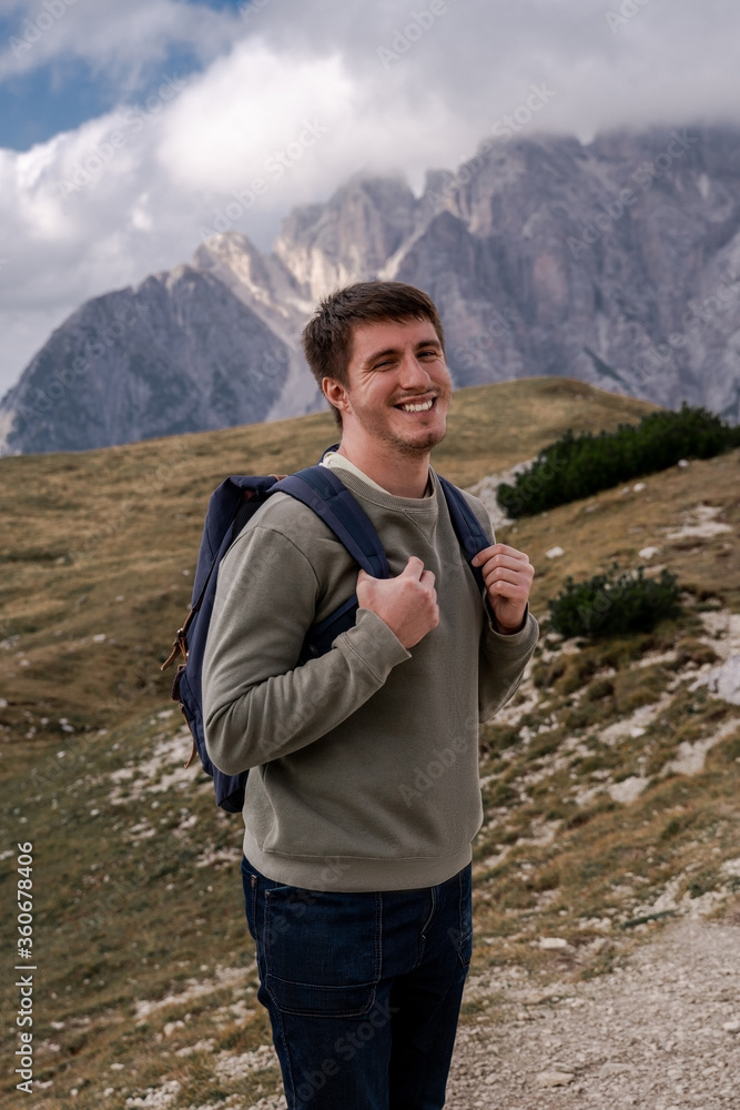 Dolomites Alps. Tre Cime di Lavaredo. Italy. Happy brunnete hiker with blue backpack smiles on alpine trail on background of rocky mountain peaks wrapped by grey clouds in summer