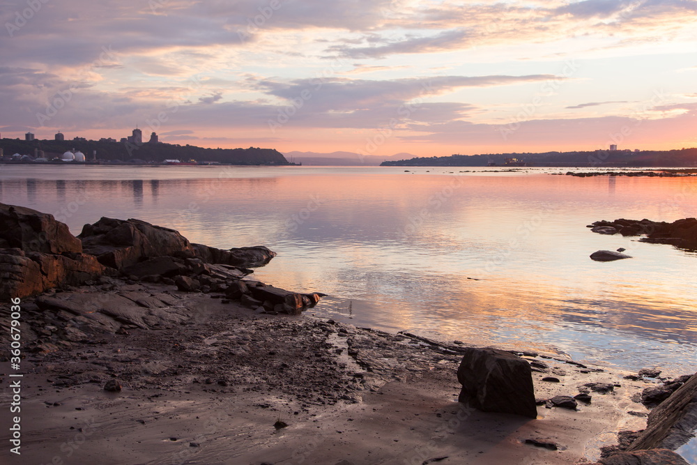 Rocky beach on the St. Lawrence River south coast with Quebec City in soft focus background seen from Lévis during a summer sunrise, Quebec, Canada