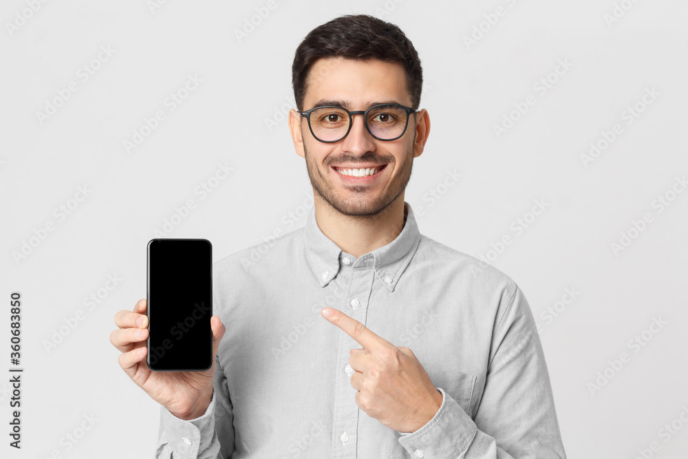Young man holding mobile phone in hand and pointing to it with finger, isolated on gray background Young man holding mobile phone in hand and pointing to it with finger, isolated on gray background