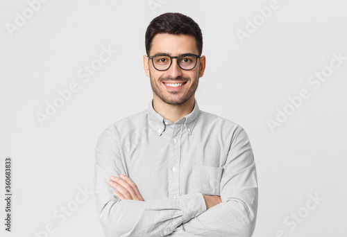 Young business guy wearing gray shirt and eyeglasses, standing with arms crossed, smiling happily and feeling confident, isolated on studio background