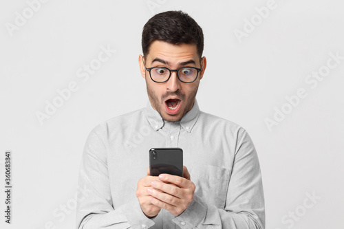 Shocked business man in casual shirt looking at his phone with surprise expression, isolated on gray background
