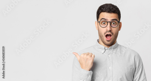 Banner of shocked young man in shirt pointing with thumb to left side, isolated on gray background with copy space photo